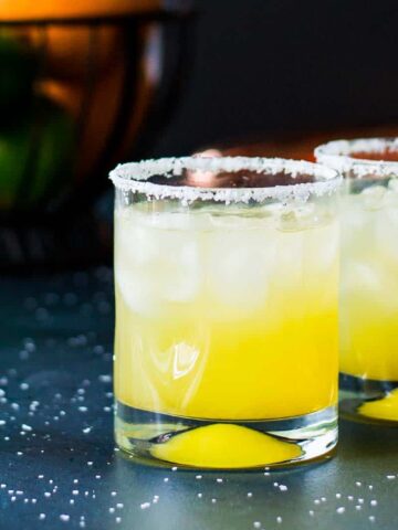 Two glasses of yellow citrus cocktail with ice and salted rims are positioned on a dark surface. A wedge of citrus rests at the bottom of each glass. In the background, a basket holds oranges and limes.