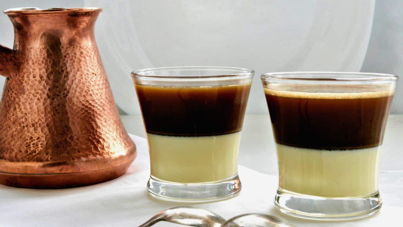 Two glasses filled with a layered beverage, featuring a creamy base topped with a dark liquid, are placed next to a copper pot on a white surface.