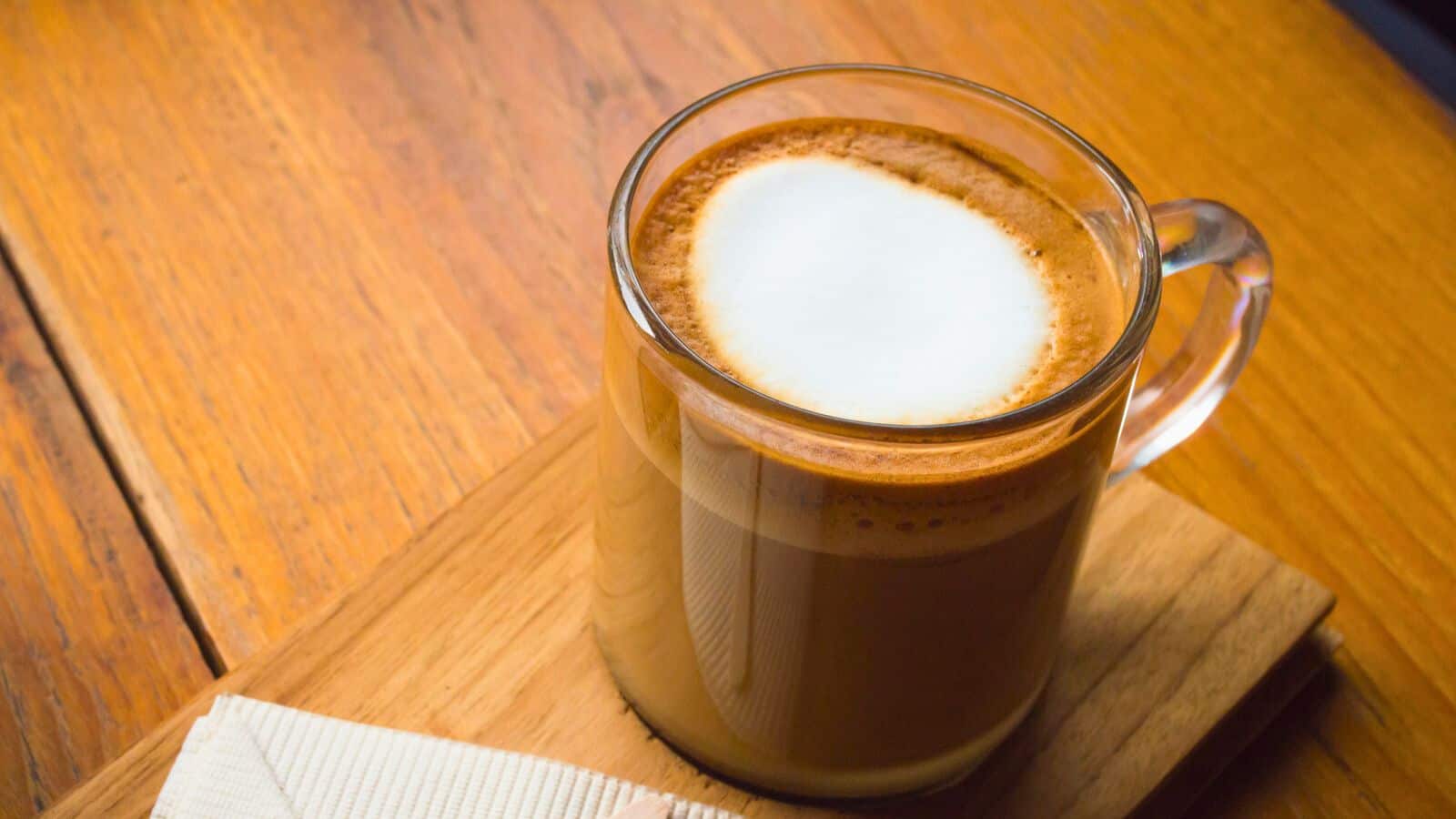 A clear glass mug filled with a foamy cappuccino sits on a wooden board. A neatly folded napkin is placed beside it. The surface is a warm-toned wooden table, and the drink has a thick layer of foam on top.