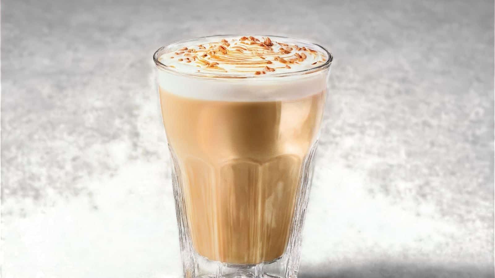 A glass of latte with frothy milk topped with a drizzle of caramel and crushed nuts, sitting on a textured gray surface.