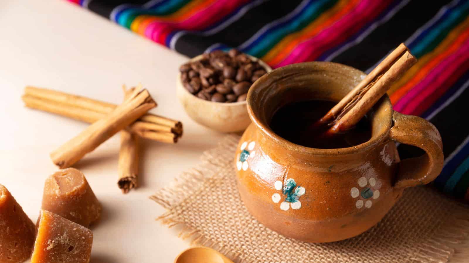 A rustic ceramic mug filled with a hot beverage and cinnamon sticks sits on a piece of burlap. Nearby are whole coffee beans in a small bowl, pieces of brown sugar, and a colorful striped fabric. A wooden spoon and more cinnamon sticks are also present.