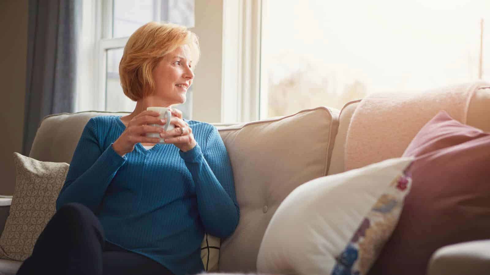 A woman with short blonde hair sits on a couch, holding a mug, looking out a window. She wears a blue sweater and is surrounded by pillows. Sunlight filters through the window, creating a warm atmosphere in the room.