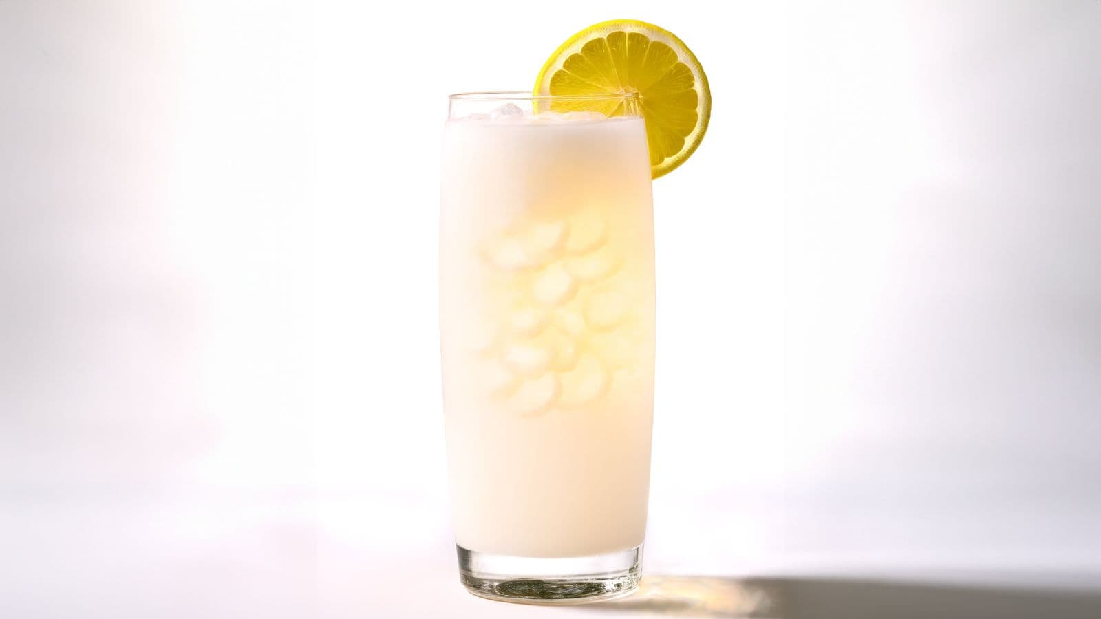 A tall glass of a creamy white beverage with ice cubes, garnished with a lemon wheel on the rim. The background is a soft white, highlighting the drink.