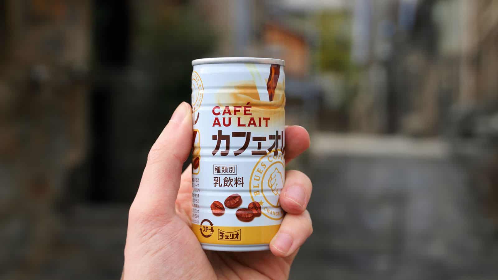 A hand holding a can of Caf&eacute; Au Lait beverage featuring coffee beans and milk swirl graphics on the label. 