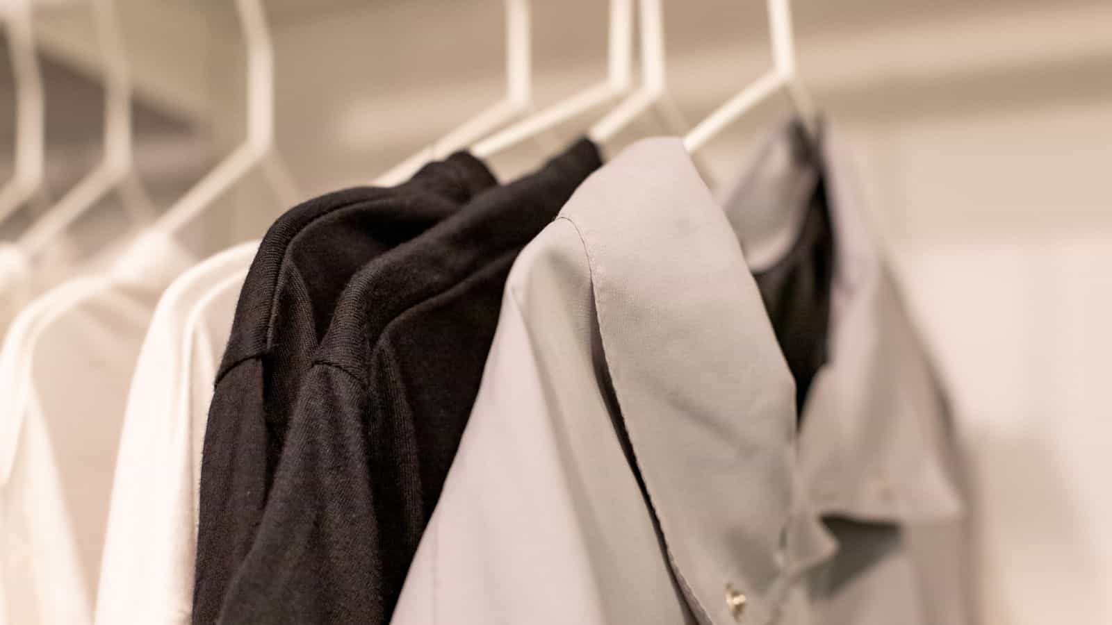 Close-up of several shirts on hangers in a closet. The shirts include a white shirt, a black shirt, and a light gray shirt. The focus is on the neatly arranged clothing hanging on white hangers.
