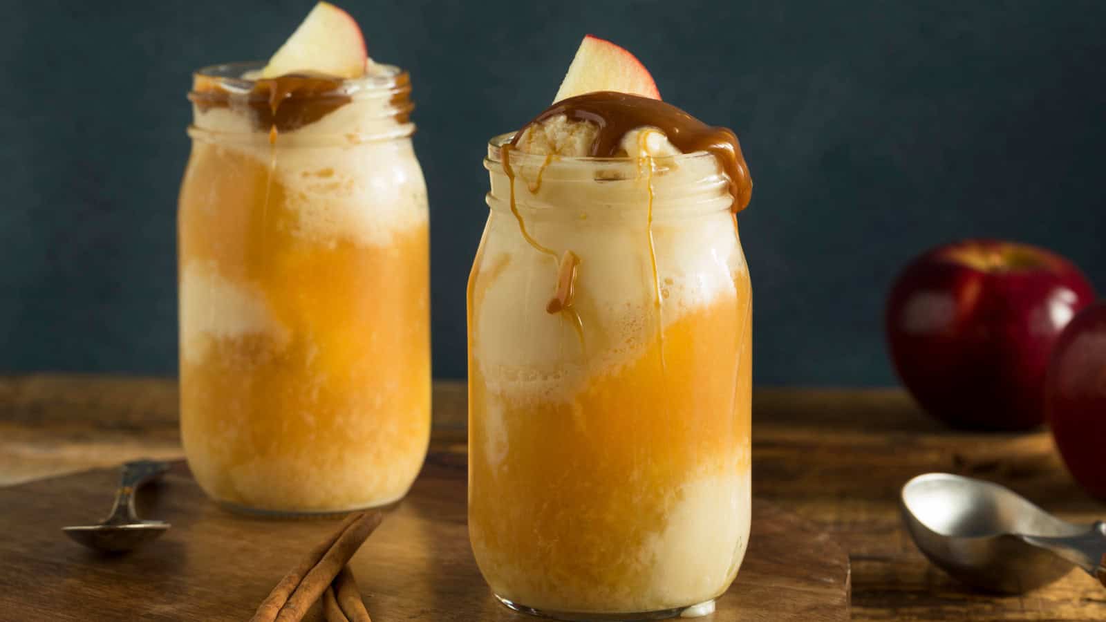 Two mason jars filled with caramel apple milkshakes, reminiscent of underrated Starbucks flavors, are topped with whipped cream and apple slices. Caramel sauce drips down the sides. A spoon and cinnamon sticks lie nearby, while apples peek from the background.