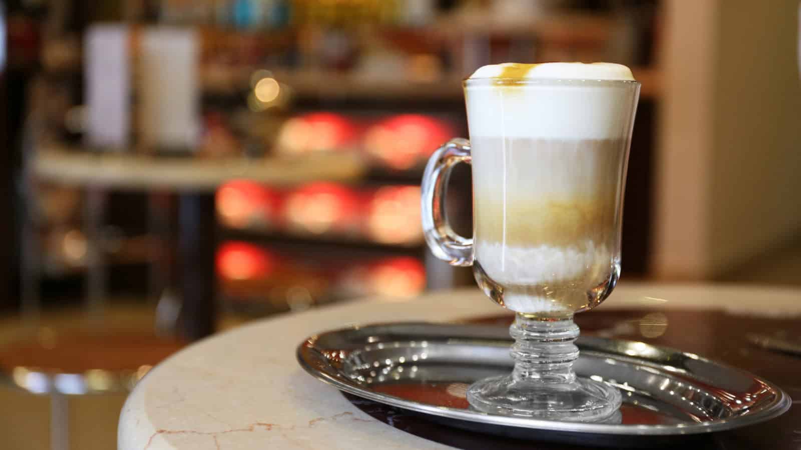A glass mug filled with a layered frothy latte sits on a silver tray. The background features blurred indoor seating and soft lighting.