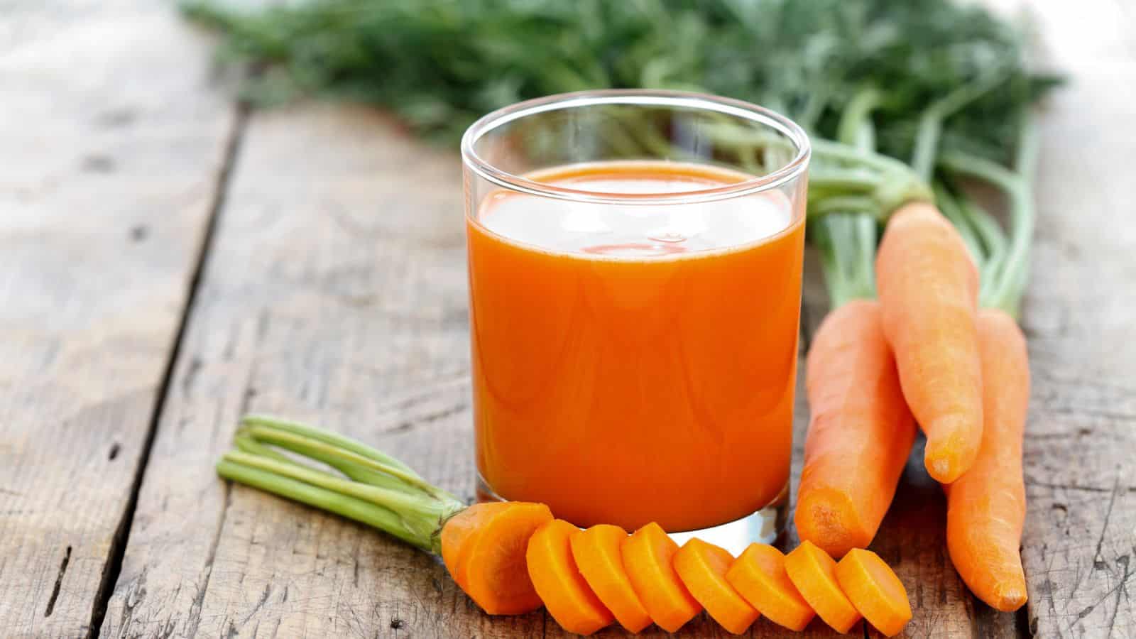 A glass of orange carrot juice stands on a wooden surface, surrounded by whole and sliced carrots. Green carrot tops are visible in the background.