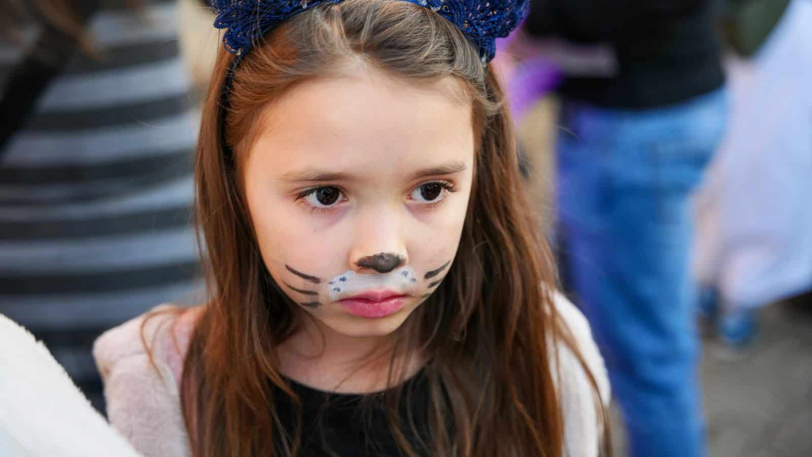 A young girl with long brown hair is wearing cat ears and has her face painted like a cat, complete with a black nose and whiskers. She appears to be in a crowd, with people and blurred motion in the background.