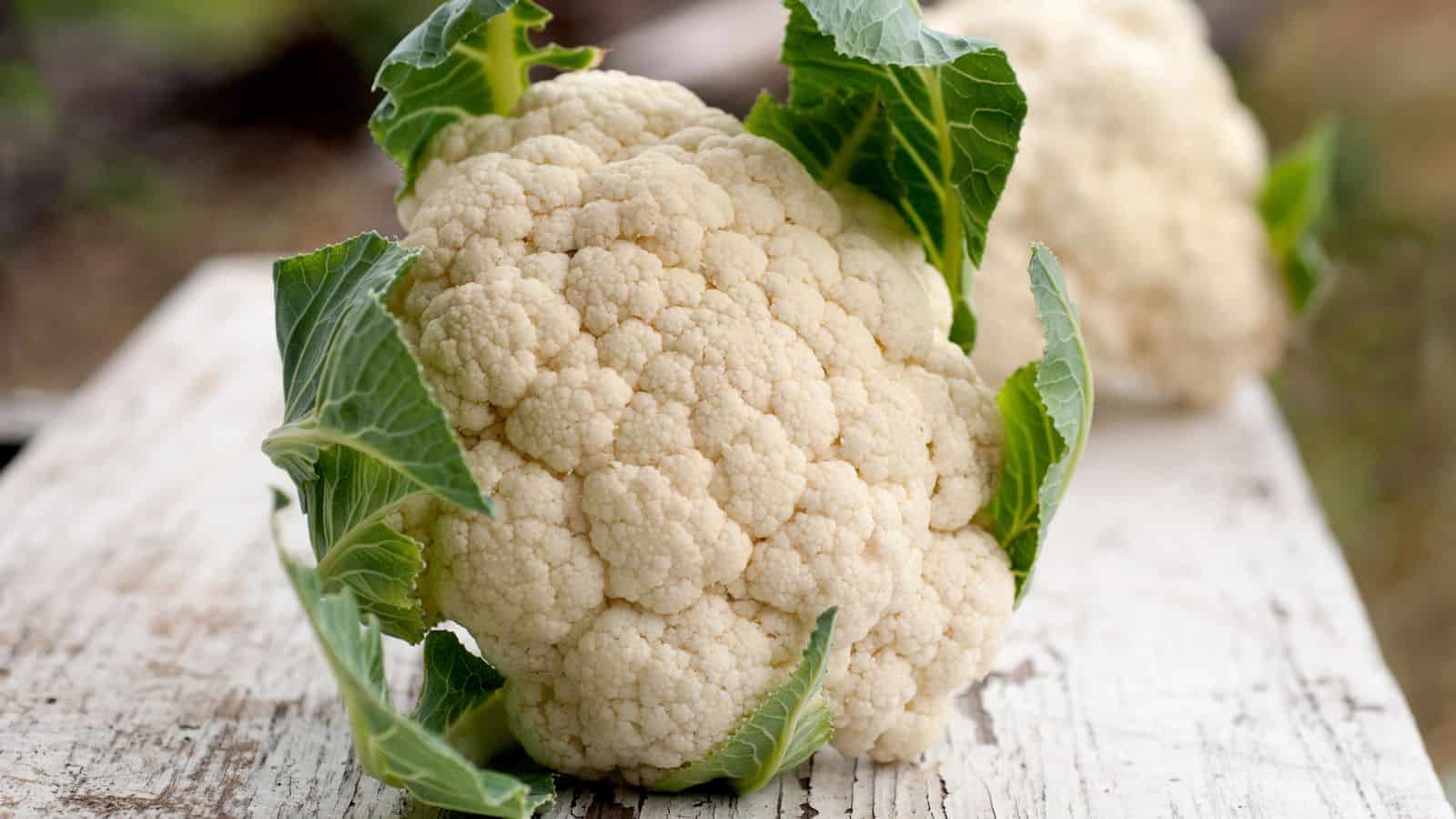 A head of cauliflower with green leaves rests on a weathered white wooden surface. Another cauliflower is blurred in the background.
