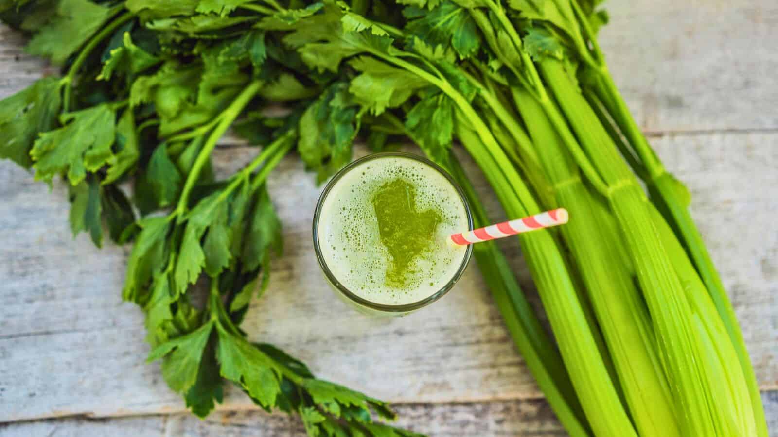 A glass of green celery juice with a red and white striped straw is placed on a textured wooden surface. Fresh celery stalks and leaves are spread beside the glass.