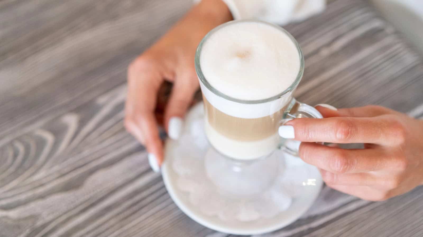 A person with manicured nails holds a glass cup of layered coffee with frothy milk on a saucer. The surface is a wood-textured table.