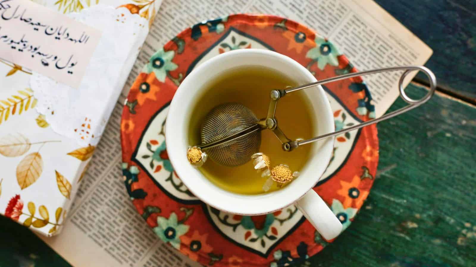 A cup of tea with a metal infuser sits on a patterned saucer, atop an open newspaper. Two small white flowers are visible near the infuser. A book with a floral cover is partially visible on the left.