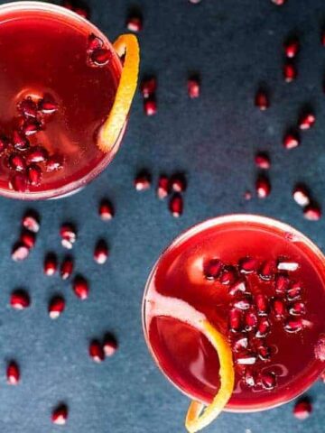 Two glasses filled with a red drink garnished with orange peel and pomegranate seeds on a dark surface. Pomegranate seeds are scattered around, and two orange slices are placed on the side.