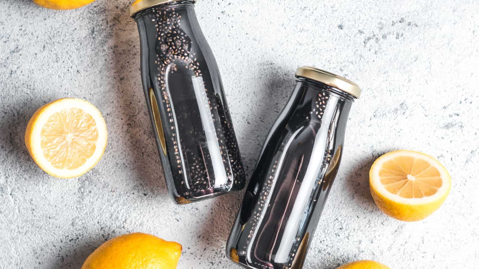 Two glass bottles filled with a dark liquid are placed on a light gray surface. Surrounding them are four lemons, two of which are halved, revealing their flesh. The scene is brightly lit, emphasizing the texture of the lemons and bottles.