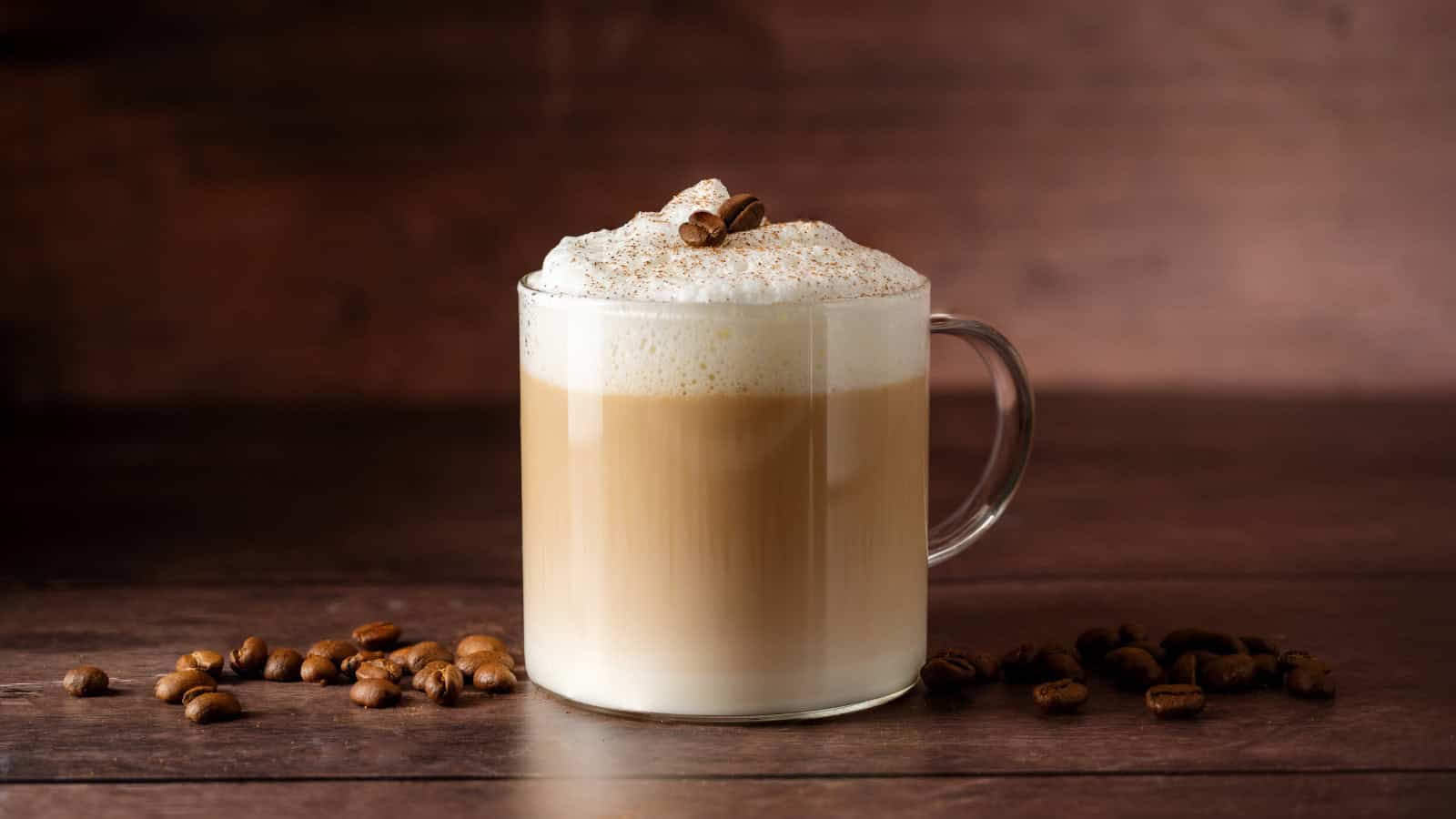 A glass mug filled with a layered coffee drink, likely a cappuccino, topped with frothy milk and garnished with chocolate shavings and coffee beans. The mug is on a wooden surface with scattered coffee beans around it.
