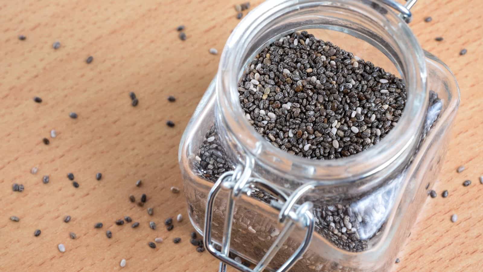 A glass jar filled with chia seeds sits open on a wooden surface. Some seeds are scattered around the jar. The jar has a metal clasp hinge lid.