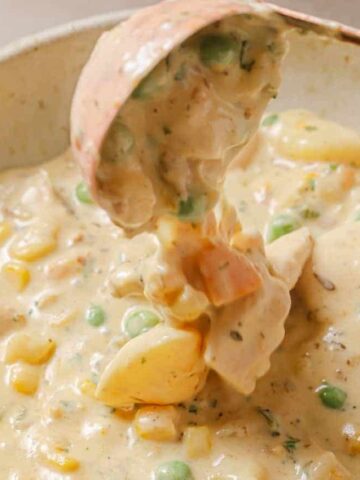 A ladle serving creamy vegetable soup is suspended above a bowl. The dish contains peas, corn, carrots, and potato chunks in a thick, beige sauce.