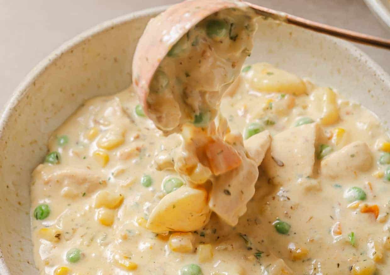 A ladle serving creamy vegetable soup is suspended above a bowl. The dish contains peas, corn, carrots, and potato chunks in a thick, beige sauce.