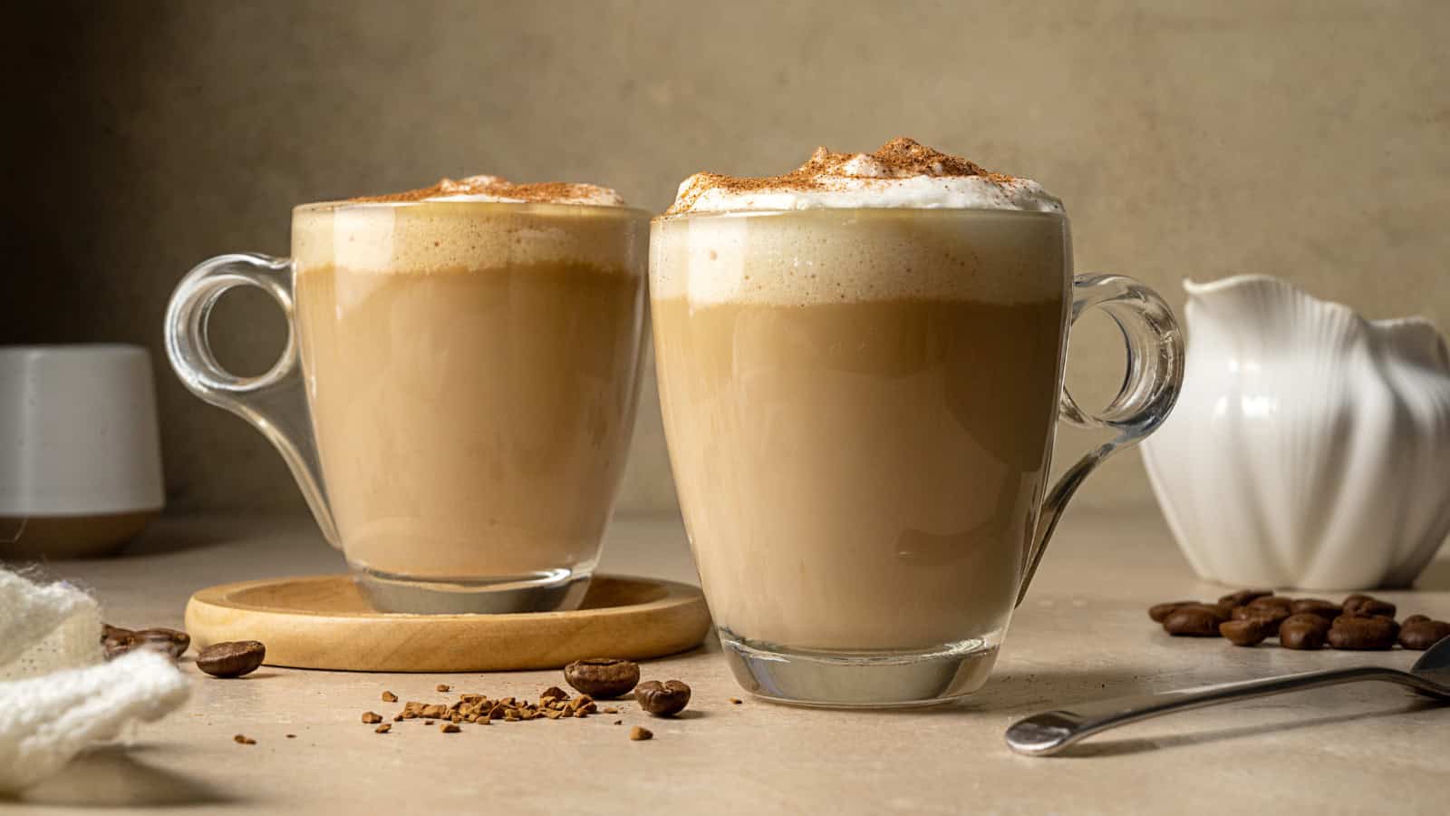 Two glass mugs filled with frothy cappuccino sit on a light-colored surface. One mug is on a wooden coaster. Coffee beans and a white ceramic creamer are nearby, with a spoon resting on the table.
