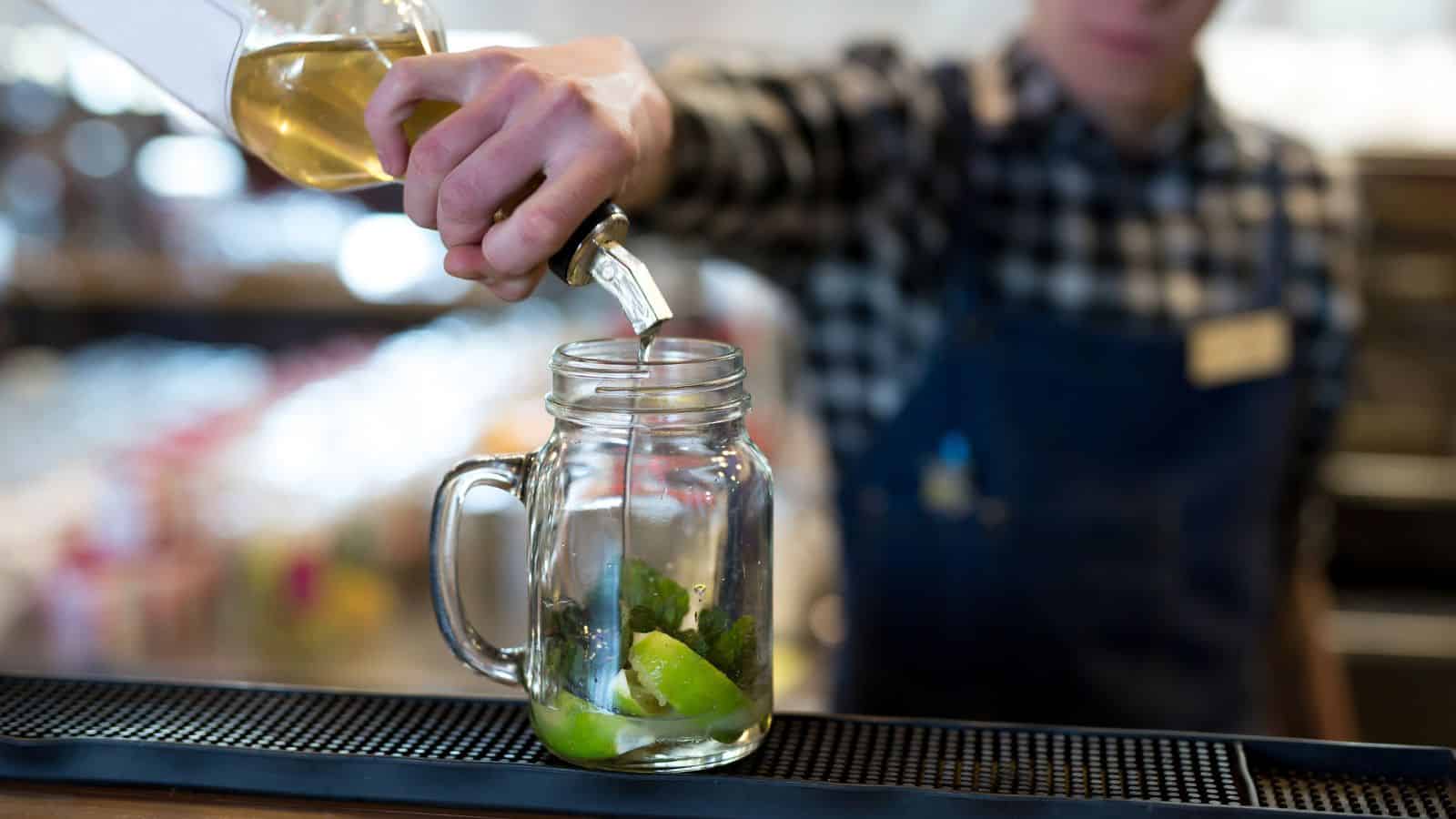 A person in a plaid shirt and denim apron pours a light-colored liquid from a bottle into a glass jar. The jar contains lime wedges and mint leaves, set on a bar counter with a blurred background.