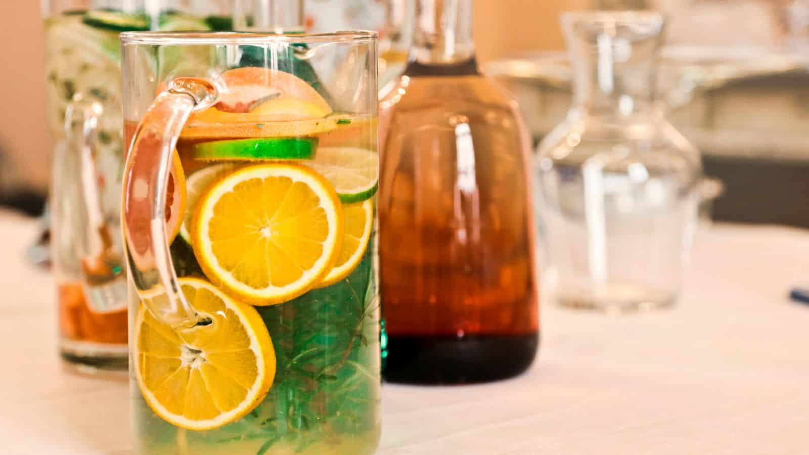 A glass pitcher filled with water, citrus slices, and cucumber on a table. Several bottles are blurred in the background.