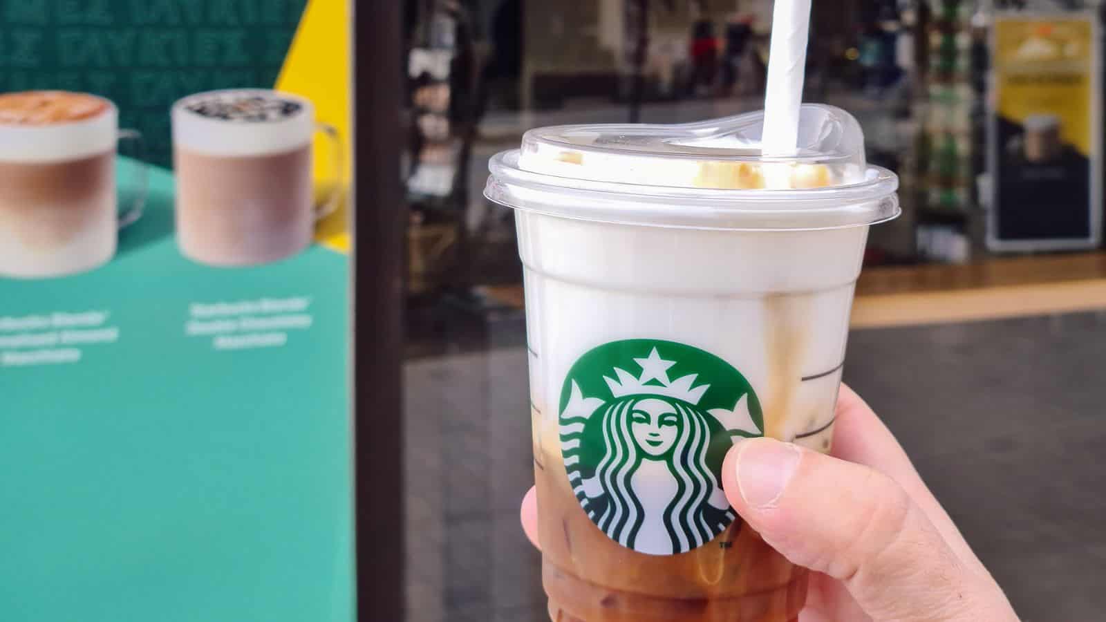 A hand holds a plastic cup of iced coffee with a straw and Starbucks logo. The drink has layers of milk and coffee. In the background, there's an outdoor Starbucks sign featuring images of two beverages.
