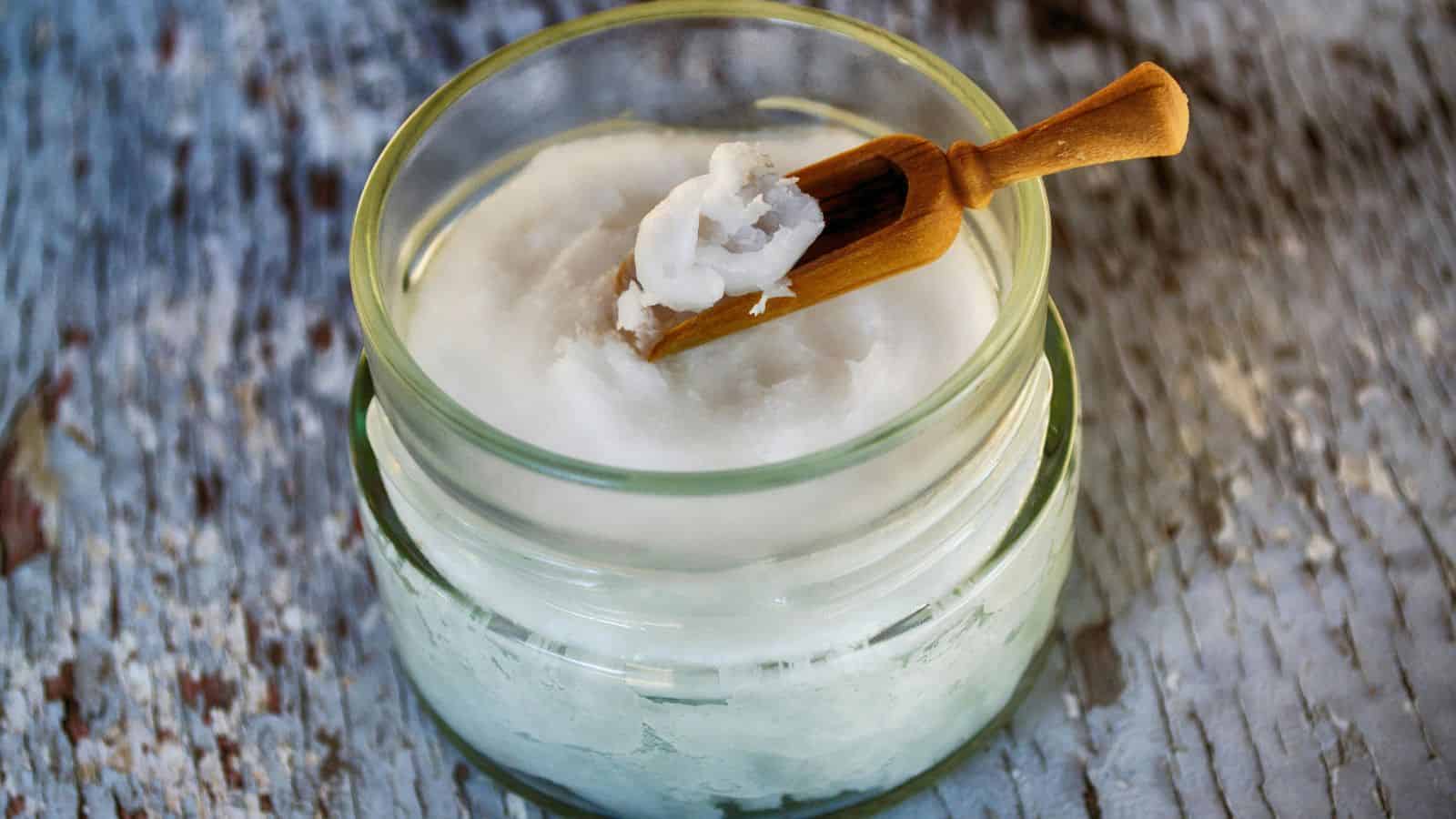 A glass jar filled with solidified coconut oil, with a small wooden spoon resting inside. The jar is placed on a textured wooden surface.