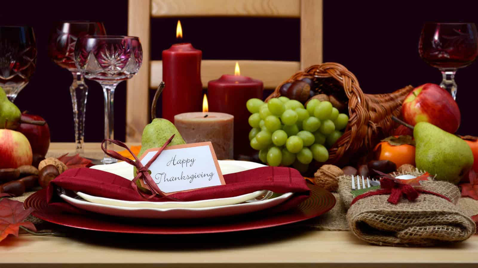 A Thanksgiving table setting with a red napkin, a note saying "Happy Thanksgiving," various candles, wine glasses, a cornucopia with fruits like grapes and pears, and decorative autumn leaves and chestnuts.