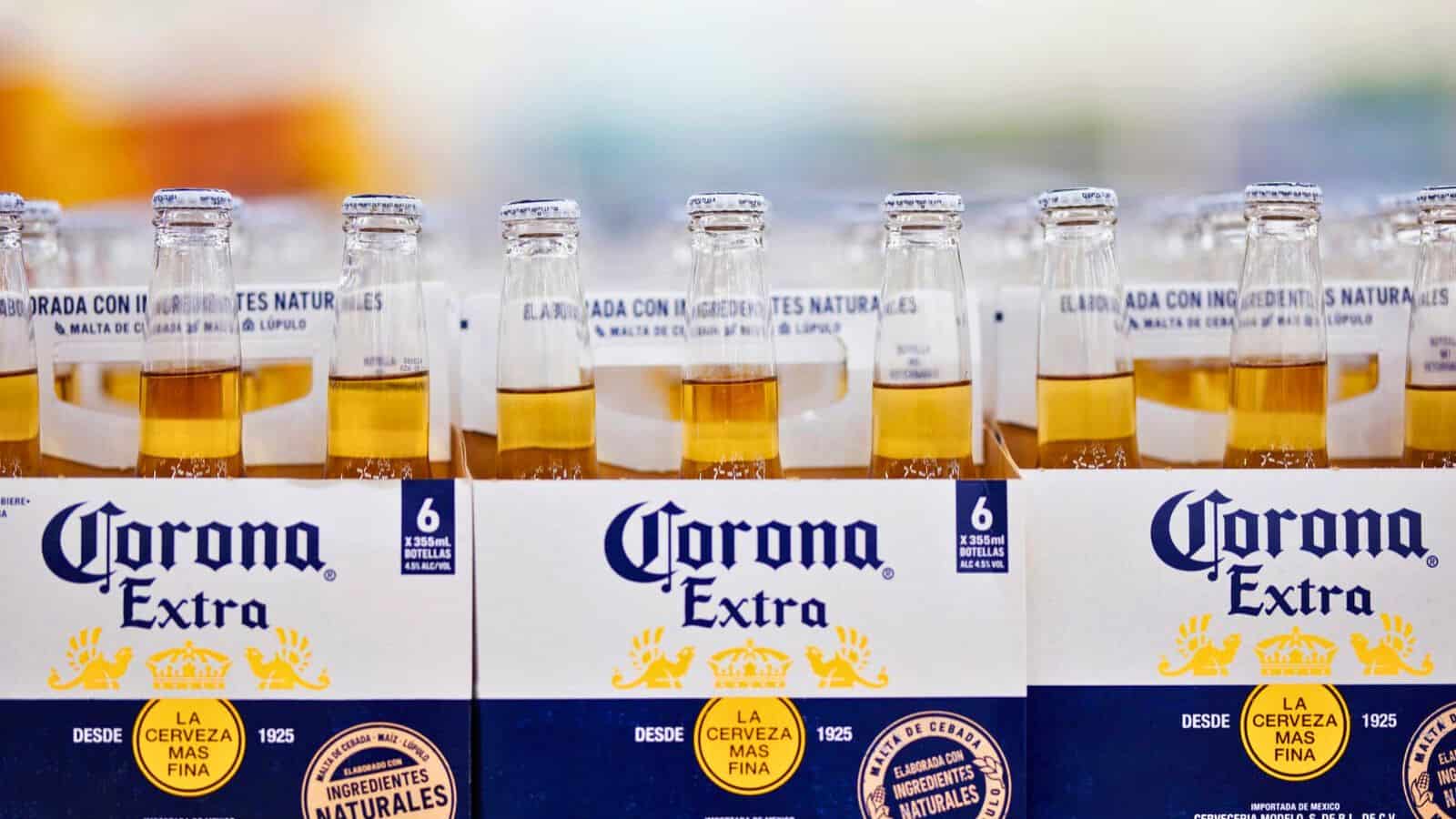A close-up of Corona Extra beer bottles arranged in a six-pack. The bottles feature a yellow liquid, and the packaging displays the brand name prominently with logos and text in Spanish.