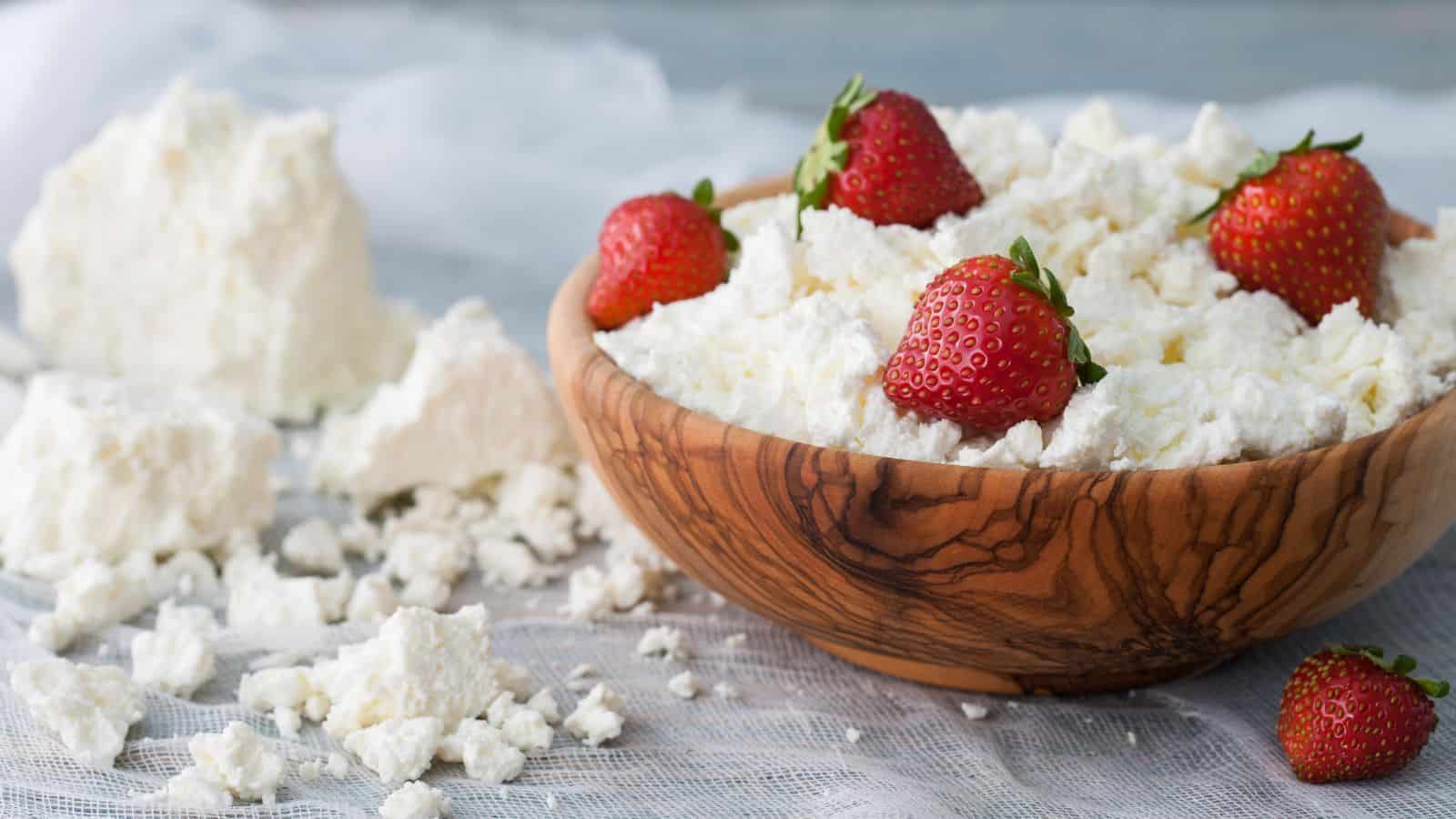 A wooden bowl filled with cottage cheese topped with fresh strawberries. More cottage cheese is scattered on a sheer white fabric in the background. Four strawberries are arranged on top of the cheese in the bowl.