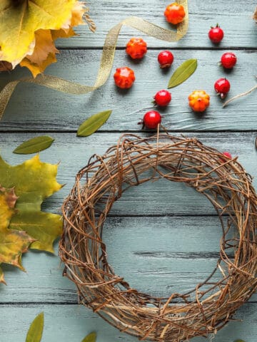 A flat lay of autumn crafting materials on a blue wooden surface. Items include a grapevine wreath, small pumpkins, yellow leaves, red berries, a spool of twine, scissors, and a ribbon.