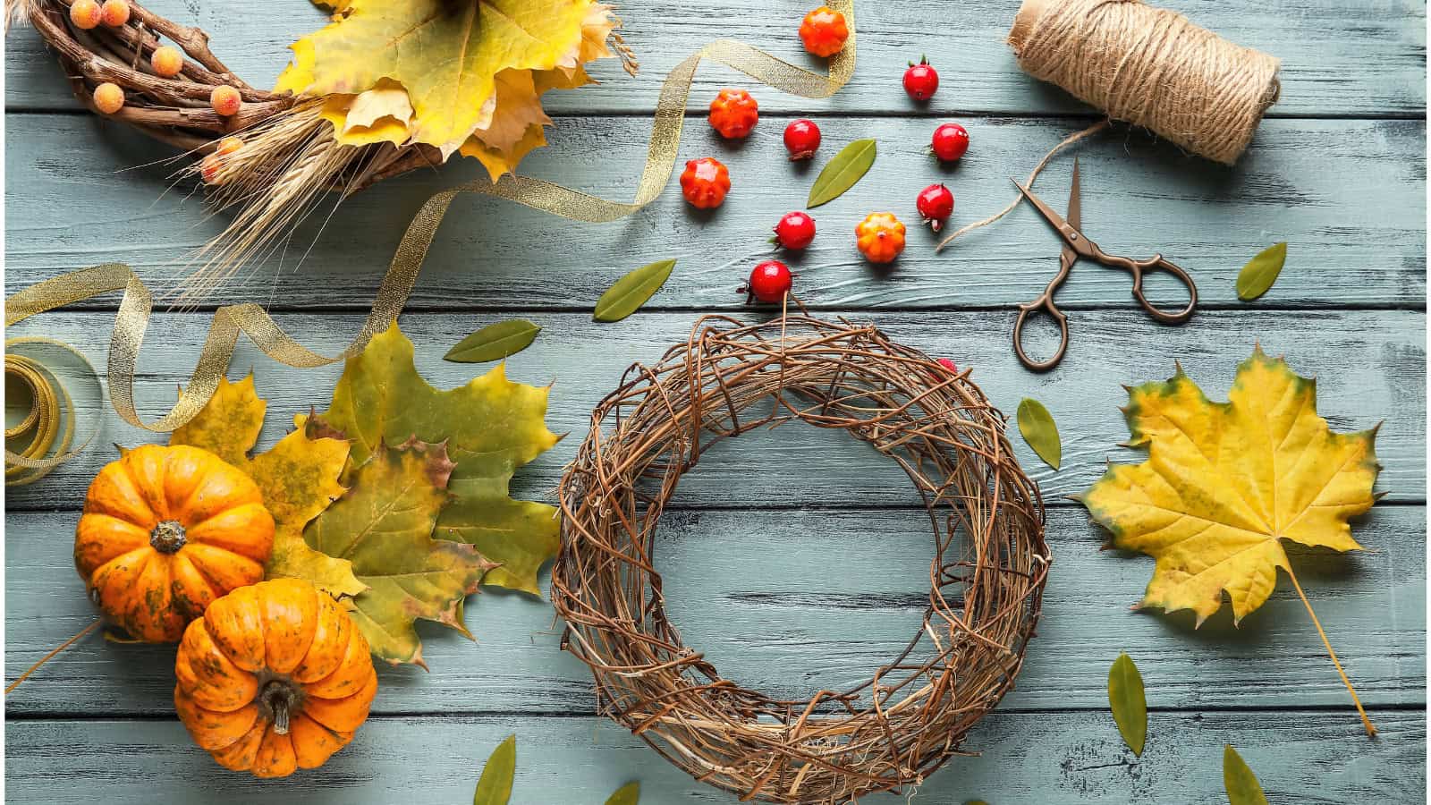 A flat lay of autumn crafting materials on a blue wooden surface. Items include a grapevine wreath, small pumpkins, yellow leaves, red berries, a spool of twine, scissors, and a ribbon.