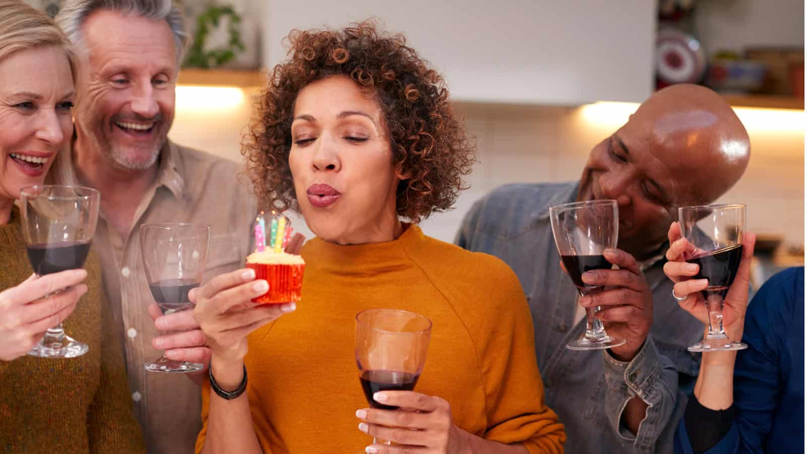 A woman in an orange sweater blows out candles on a cupcake while holding a glass of wine. Four people stand around her smiling, each holding a glass of red wine. The setting appears to be a kitchen.