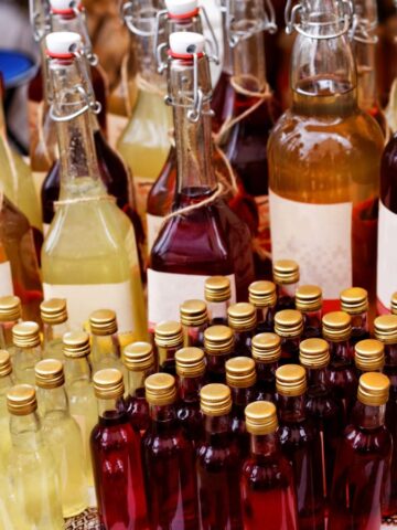 An assortment of bottles filled with various colored liquids is displayed on a table. The bottles have different shapes and sizes, with some sealed by corks and others by swing tops. A patterned cloth is partially visible underneath.