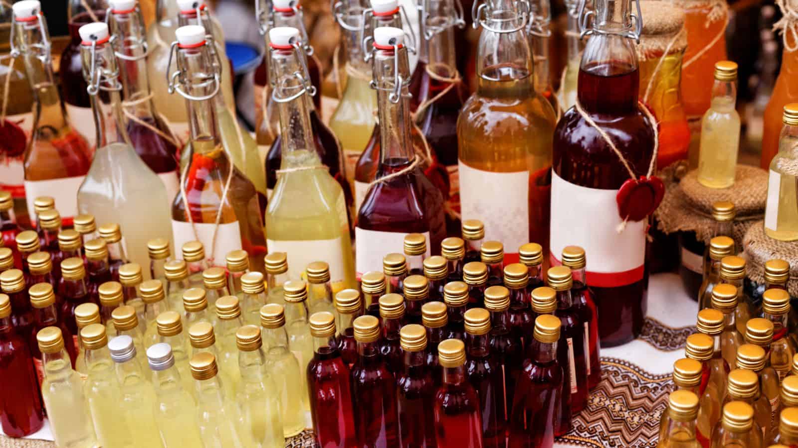 An assortment of bottles filled with various colored liquids is displayed on a table. The bottles have different shapes and sizes, with some sealed by corks and others by swing tops. A patterned cloth is partially visible underneath.