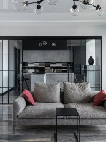 A modern living room with a gray sofa featuring two red cushions, a small black side table, and glass doors leading to the kitchen. The floor is glossy, and there is a black ceiling light fixture with round bulbs.