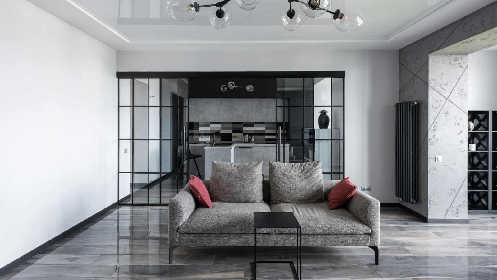 A modern living room with a gray sofa featuring two red cushions, a small black side table, and glass doors leading to the kitchen. The floor is glossy, and there is a black ceiling light fixture with round bulbs.