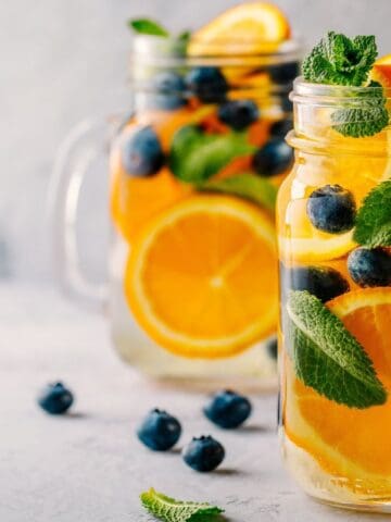 Two mason jars filled with a refreshing drink, featuring water infused with orange slices, blueberries, and mint leaves. Additional fruits and mint leaves are scattered around the jars on a light surface.