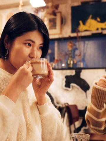 Two people are sitting in a cozy cafe, sipping drinks from glass mugs. The person on the left is wearing a white fleece top, and the one on the right is wearing a striped sweater. The background has various art pieces on the walls.