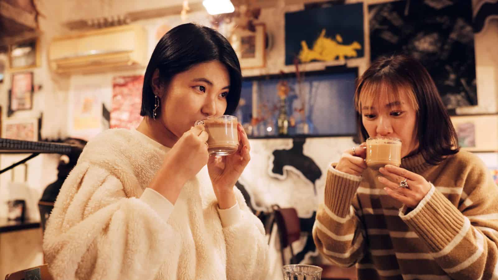 Two people are sitting in a cozy cafe, sipping drinks from glass mugs. The person on the left is wearing a white fleece top, and the one on the right is wearing a striped sweater. The background has various art pieces on the walls.