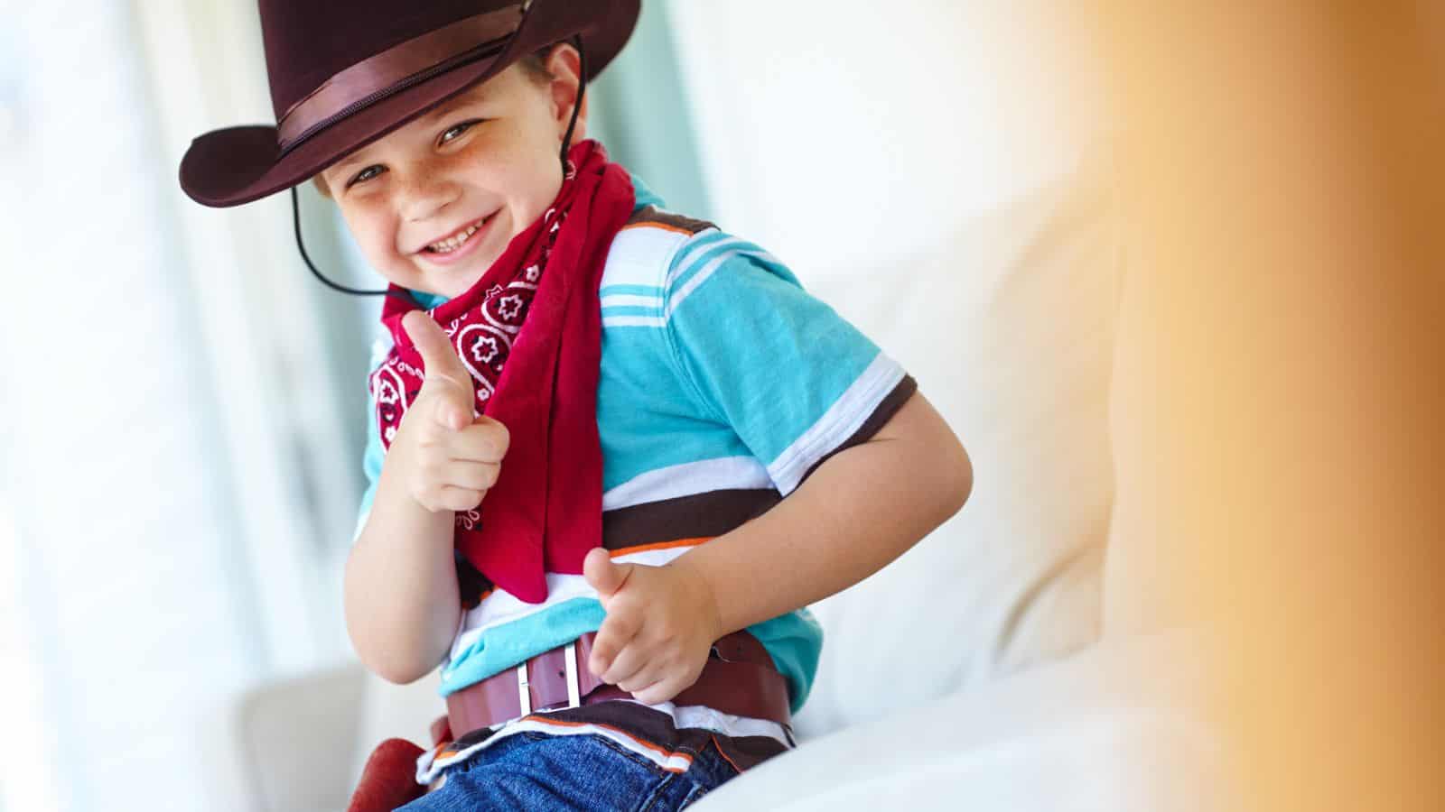 A child wearing a cowboy hat, red bandana, and blue shirt points finger guns while smiling. The child sits on a light-colored sofa indoors.