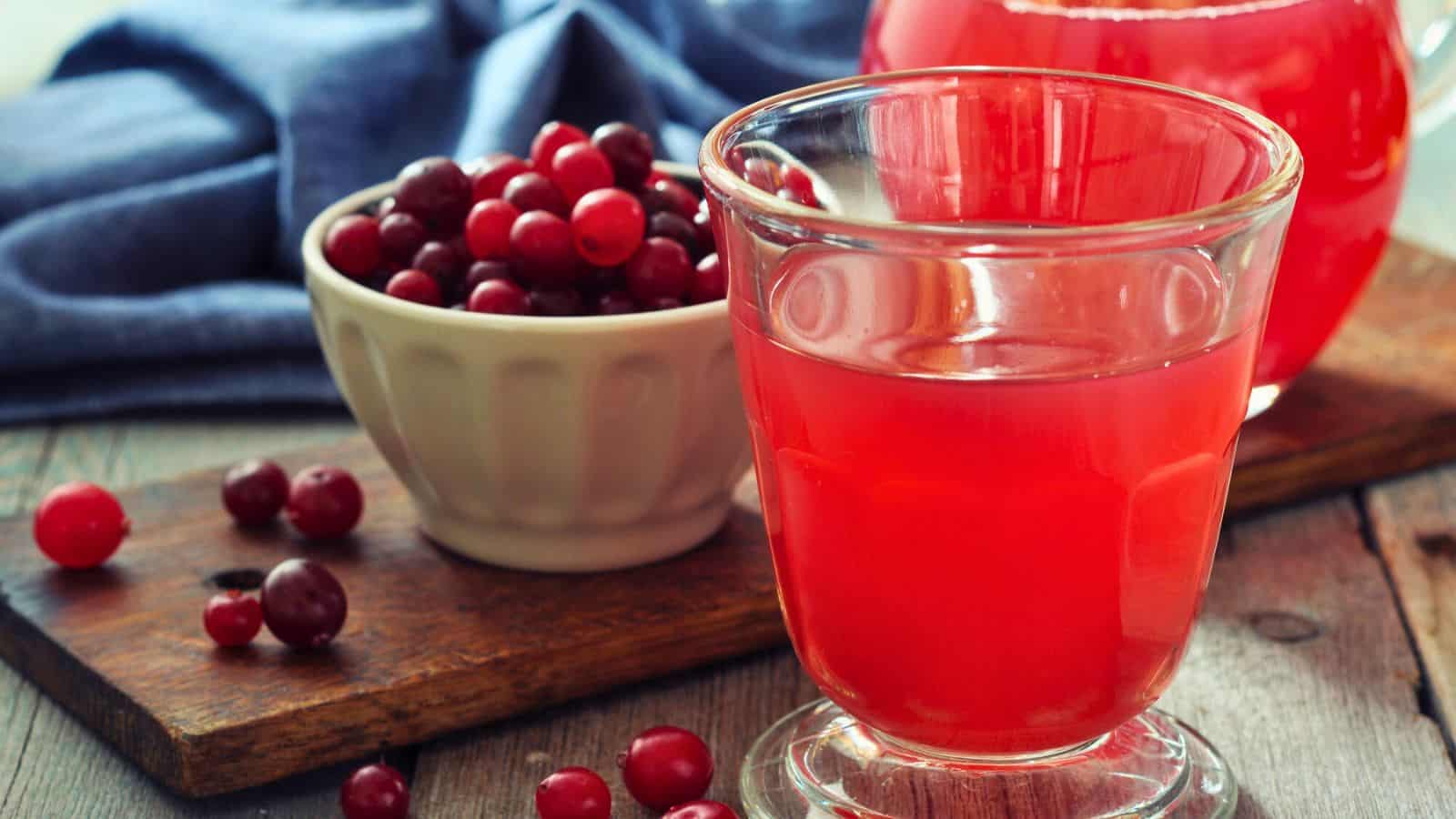 A glass of cranberry juice is on a wooden surface next to a white bowl filled with fresh cranberries. A larger container of juice and scattered cranberries are in the background, alongside a blue cloth.