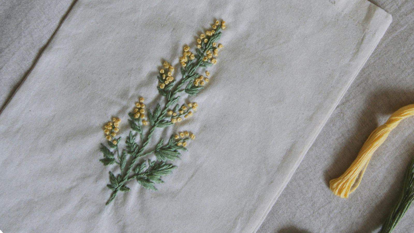 A piece of fabric featuring an embroidered design of a plant with yellow flowers and green leaves. A skein of yellow thread lies nearby on the right.
