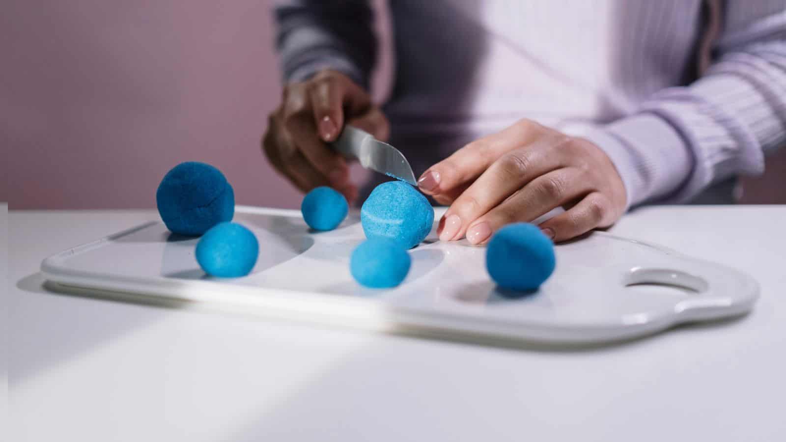 A person slices a blue foam ball with a knife on a white cutting board. Several other blue foam balls of varying sizes are scattered around the board. The person is wearing a long-sleeved top.