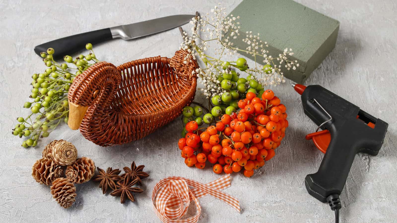 A flat lay of crafting materials includes a wicker basket, a knife, floral foam, a glue gun, clusters of orange and green berries, dried flowers, star anise, pine cones, and an orange checked ribbon on a textured surface.