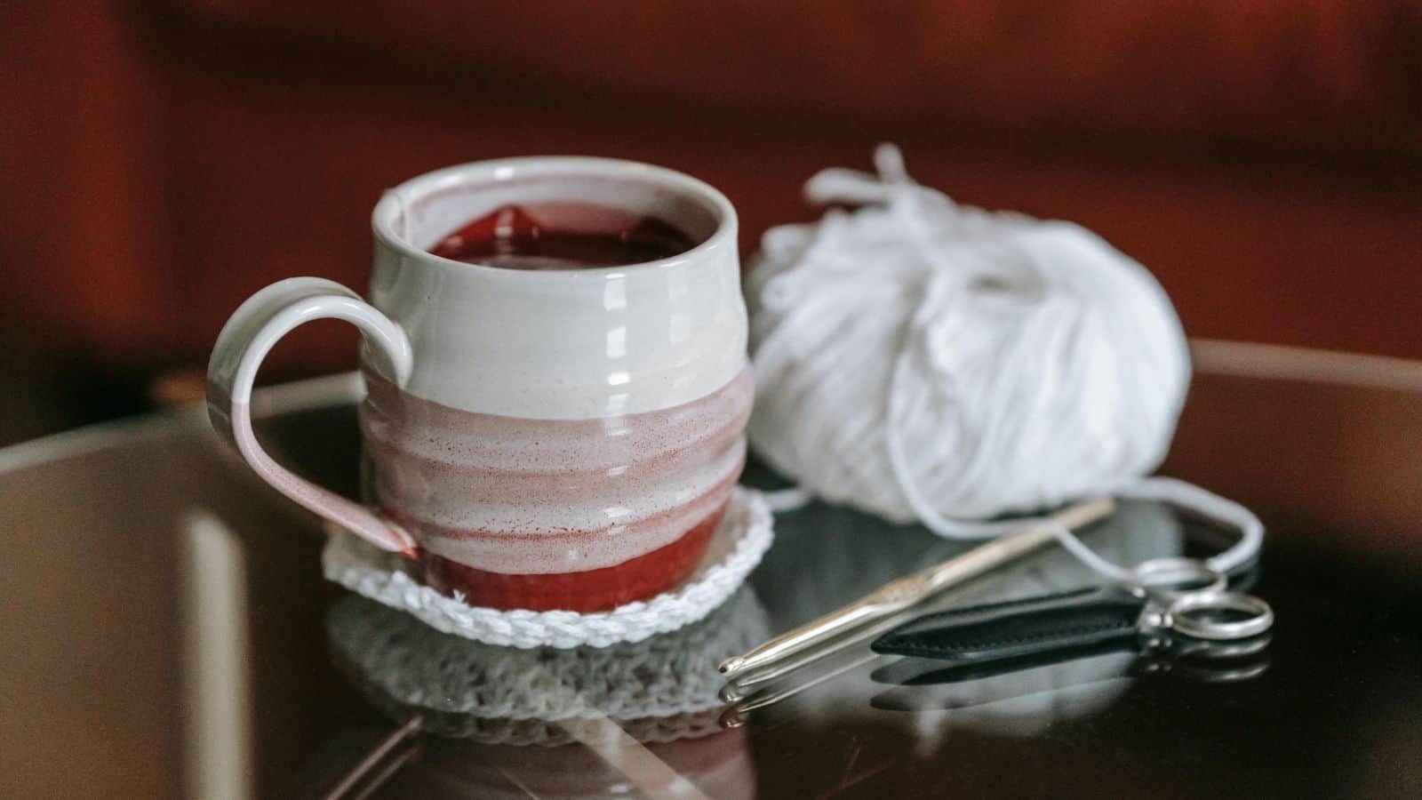 A ceramic mug containing a red beverage sits on a glass table with a crochet coaster. Beside it, there is a ball of white yarn, a crochet hook, and a pair of scissors with a black handle.