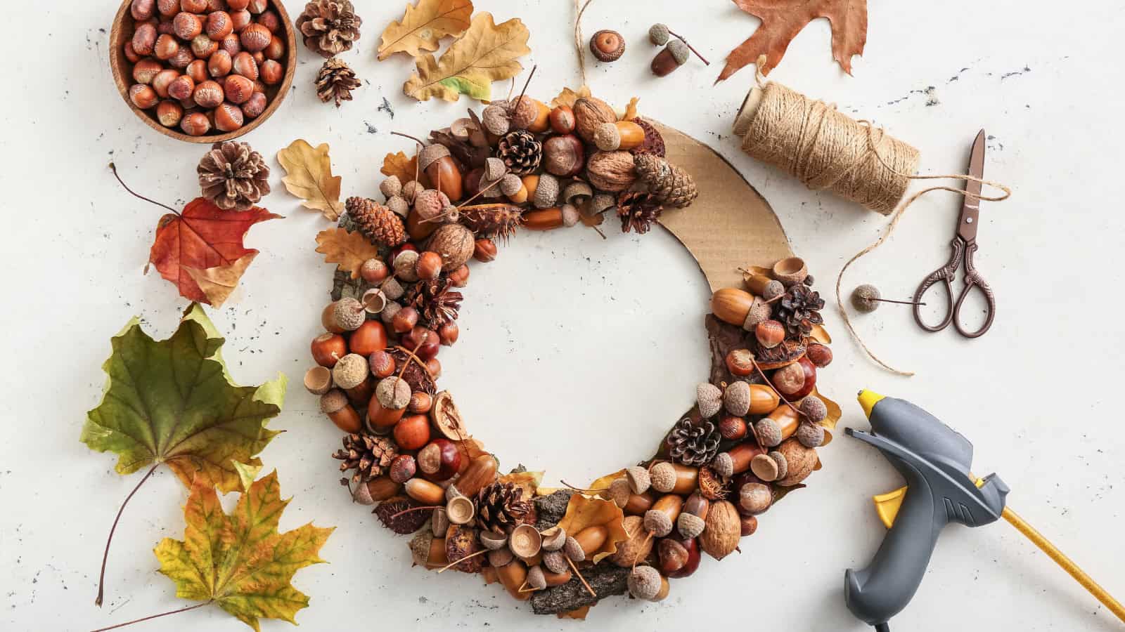A DIY autumn wreath in progress on a table. The wreath is adorned with nuts, acorns, pinecones, and leaves. Surrounding items include twine, scissors, a hot glue gun, loose leaves, and a bowl of nuts.