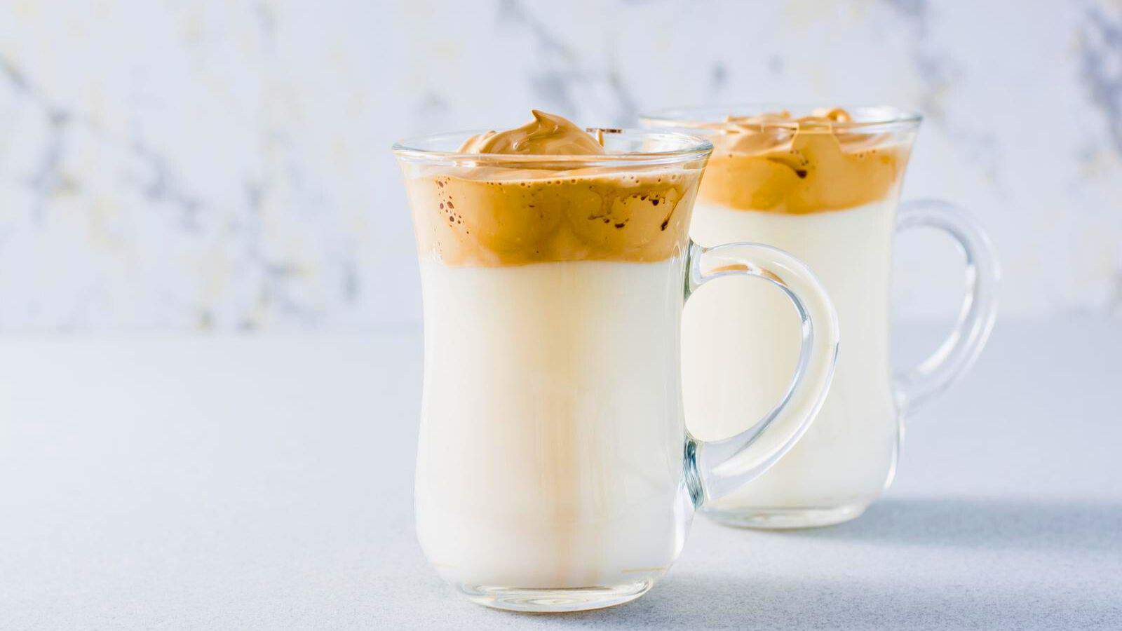 Two clear glass mugs are filled with a creamy white liquid and topped with a thick layer of whipped coffee. They are set on a light gray surface against a blurred light background.