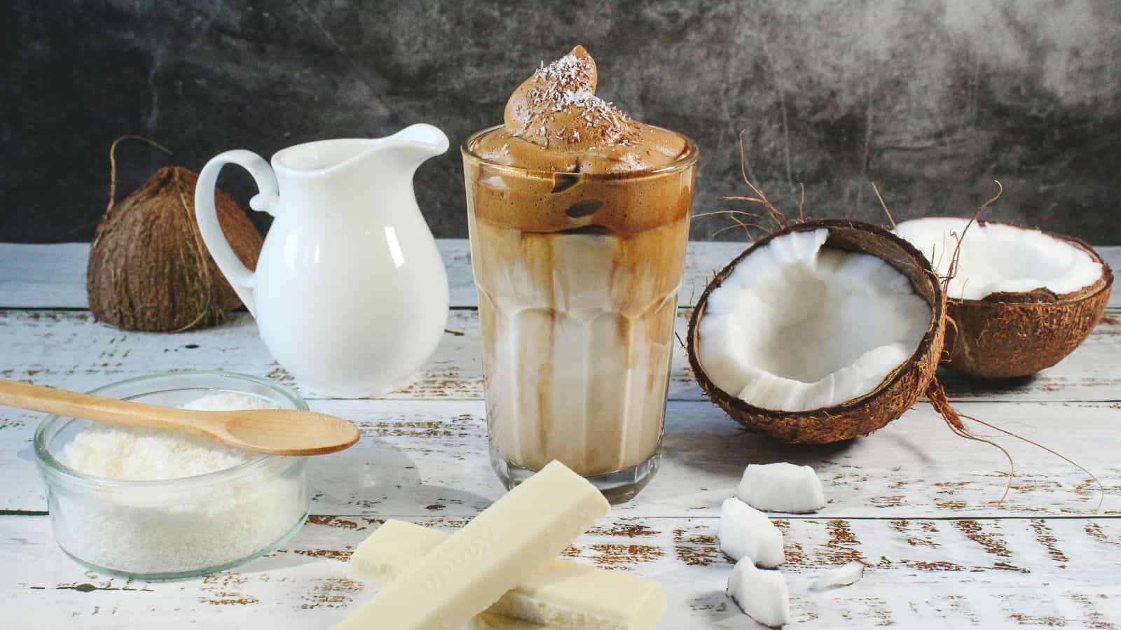 Iced coffee with whipped topping in a glass, surrounded by a jug, halved coconuts, grated coconut in a bowl with a wooden spoon, and pieces of white chocolate on a rustic white surface.